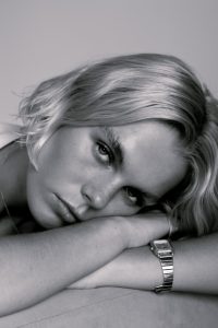 a blonde white female laying on her arms looking into the camera with no particular expression. she is wearing a silver watch that is visible and her hair falls on her face and arms naturally