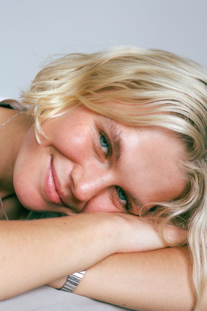 This image is part of the portfolio. In this image there is a white female smiling at the camera with her head rested on her arms. she has blonde short hair that reaches the jaw of face. This image is part of my portfolio that showcases my skills.