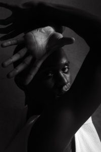 This image is part of my portfolio showcases natural light photography and creating art with posing. In this image a black and white image of a black male looking into the camera with his hands creating an interesting shape that covers parts of his face. he wears a simple white vest and no jewellery.