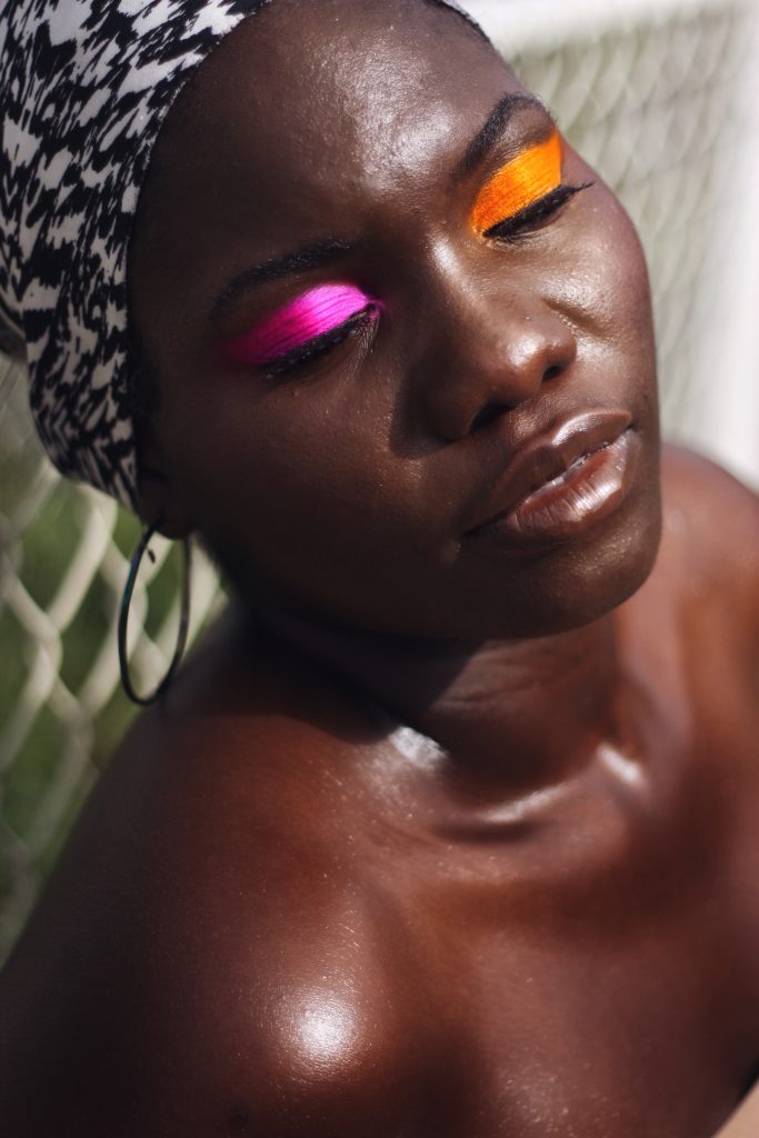 This image is part of the portfolio which comes from the editorial project I produce whilst visiting family. In this image there is a black female wearing colourful eyeshadow with one have pink on the right eye and orange on the left. Her eyes are closed in the photo as she gives a relaxed expression whilst posing in front of a white metal fence. She also wears a scarf that has a white black an d pink design.