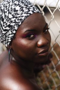 The image is from an editorial photoshoot with beautiful lady of darker complexion with vibrant colourful makeup and a headscarf on. She is posing in front of a fence with a fierce or serious express whilst looking the direction of the camera.